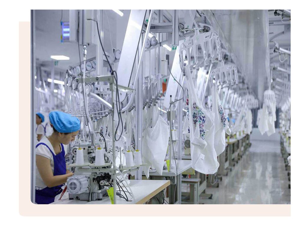 Worker operating a sewing machine in a modern garment factory with automated equipment and hanging white garments.