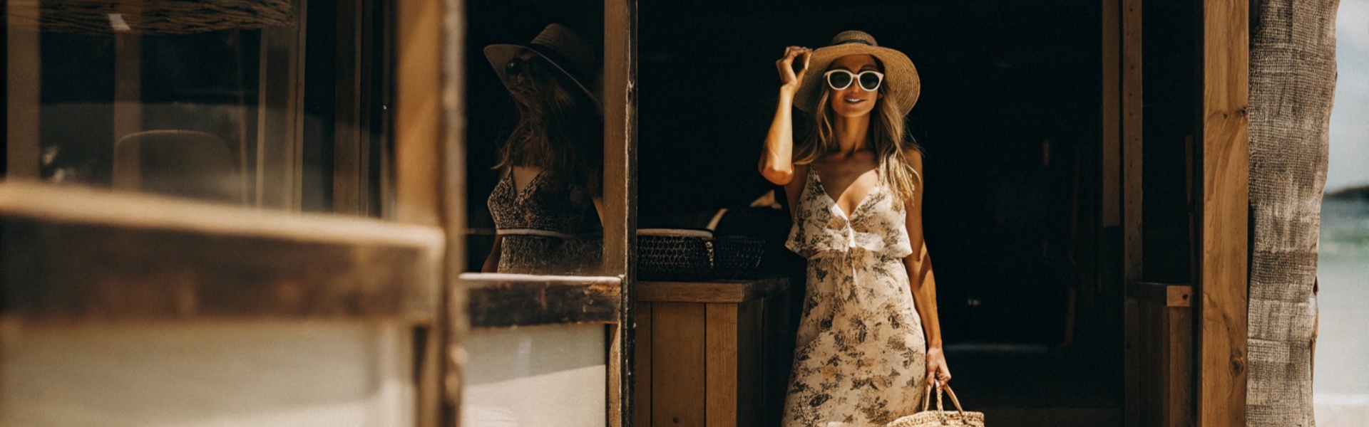 Woman in a floral maxi dress with sunglasses and a straw hat, holding a woven bag, stepping out of a wooden beach hut.