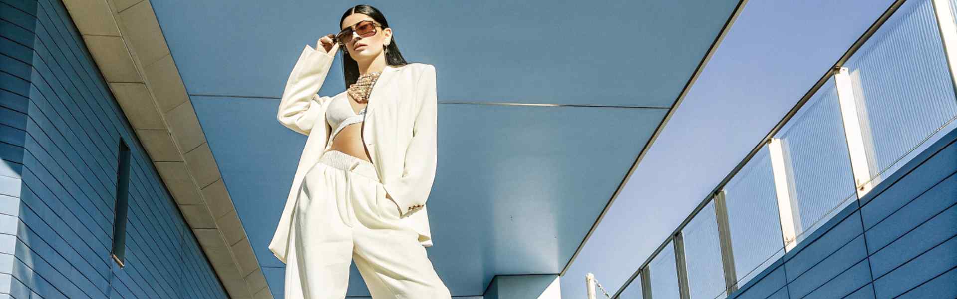 Woman in white suit with bralette posing under a modern blue structure.