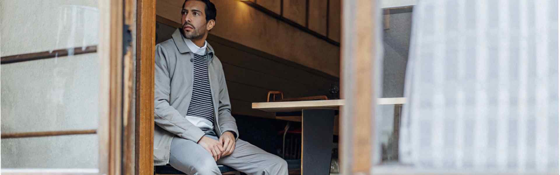 A man dressed in a gray outfit, sitting by a café entrance, wearing white sneakers.