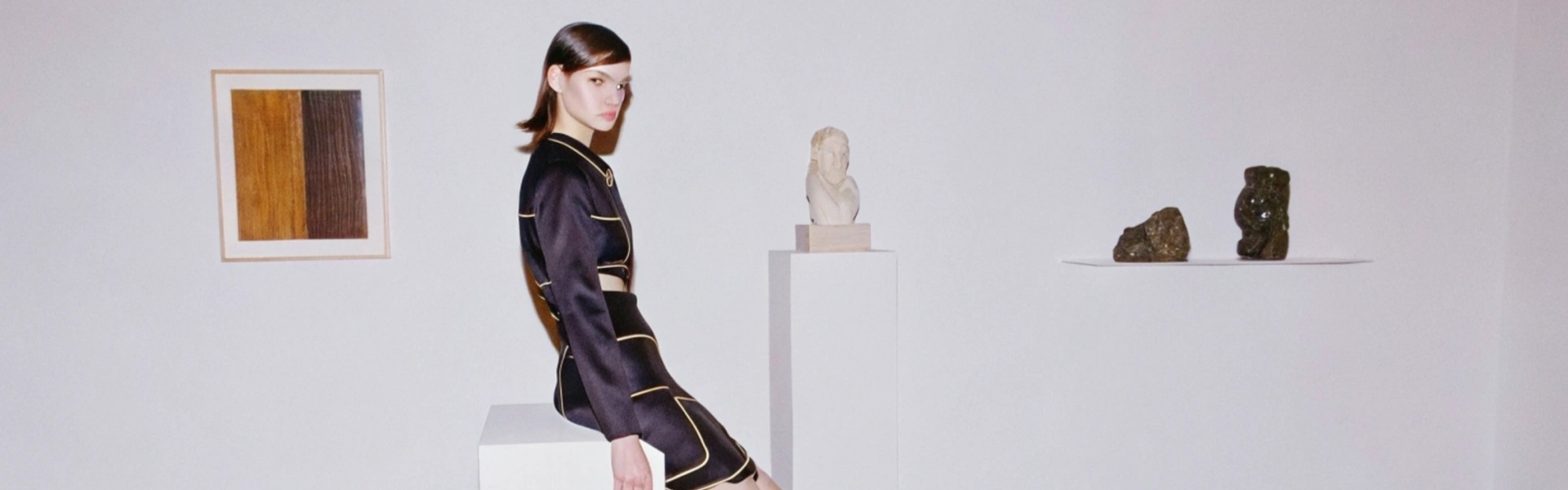 Woman in a black dress with gold accents sitting on a white pedestal in a minimalist art gallery.