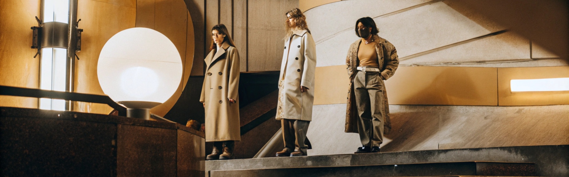 Three women in stylish neutral-toned coats and contemporary outfits stand on stairs in a modern architectural space with dramatic lighting.