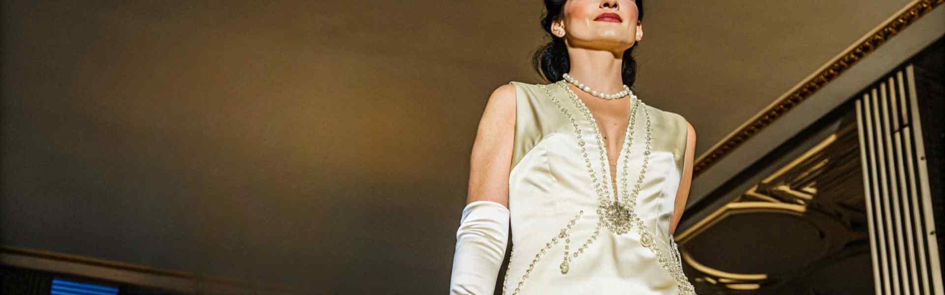 Woman in an ivory satin gown with pearl details, standing under a chandelier.