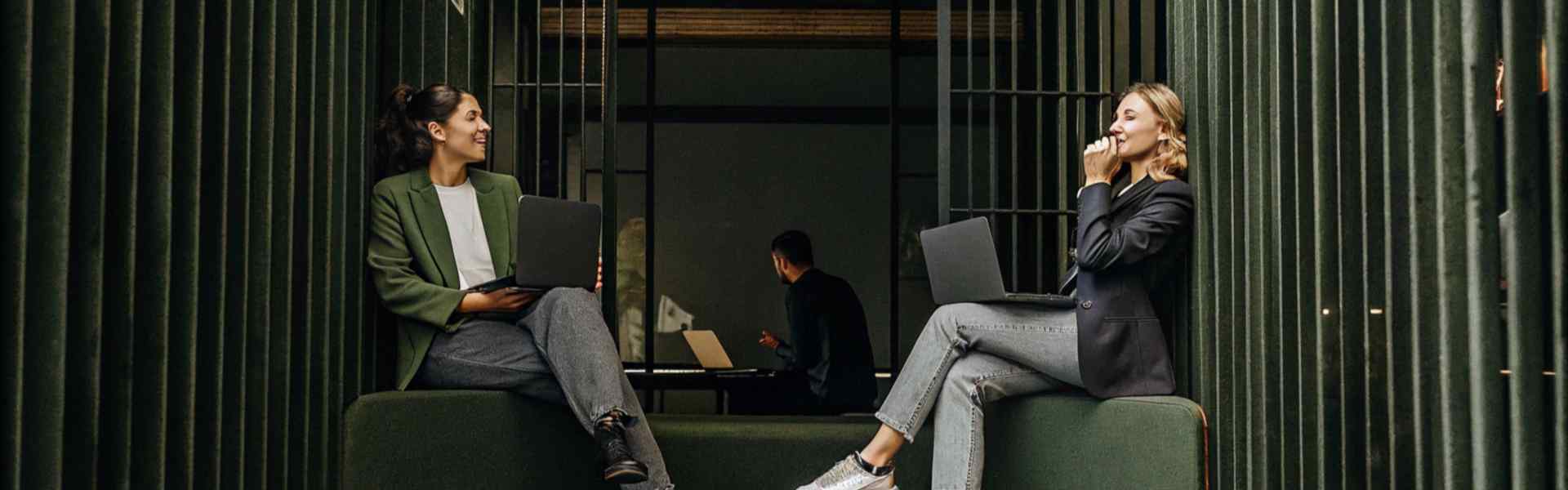 Two women sitting on green seats with laptops in a modern office.