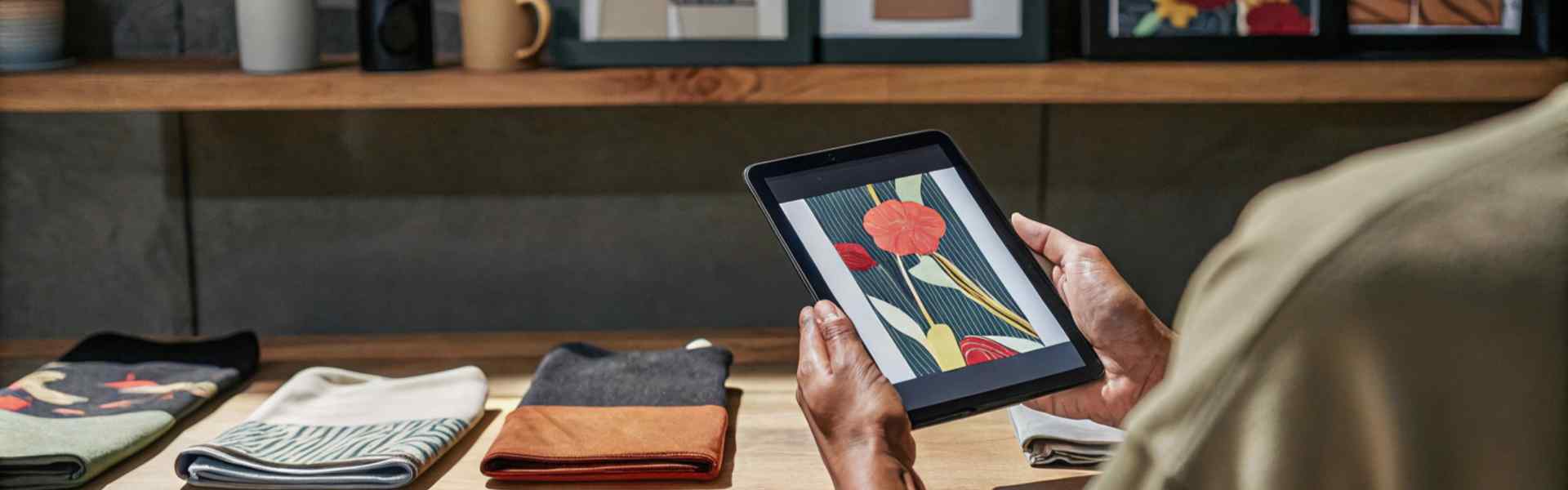 A person using a tablet to view a floral fabric design, with neatly folded scarves and art prints displayed on the table.