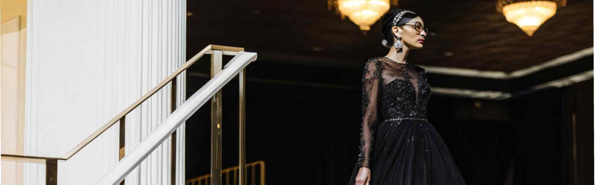 Woman in a sparkling black evening gown on a white staircase under warm chandelier lights.