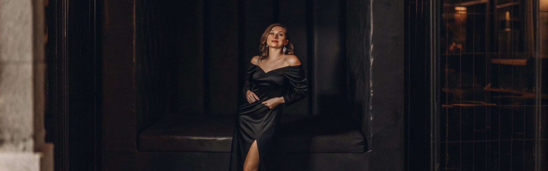 Woman in a sleek black evening gown posing under string lights.