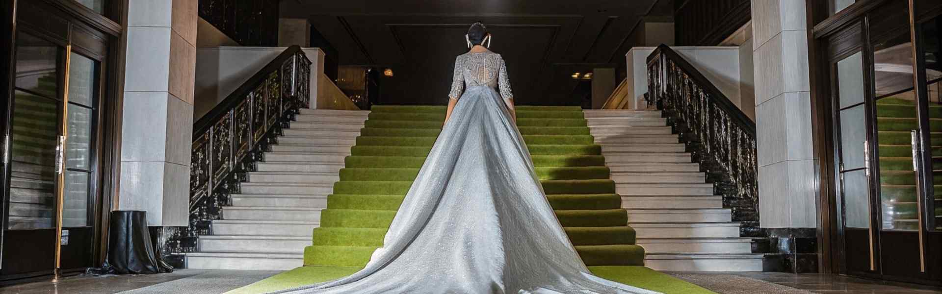 Woman in a long silver gown walking up a green-carpeted staircase beneath chandeliers.