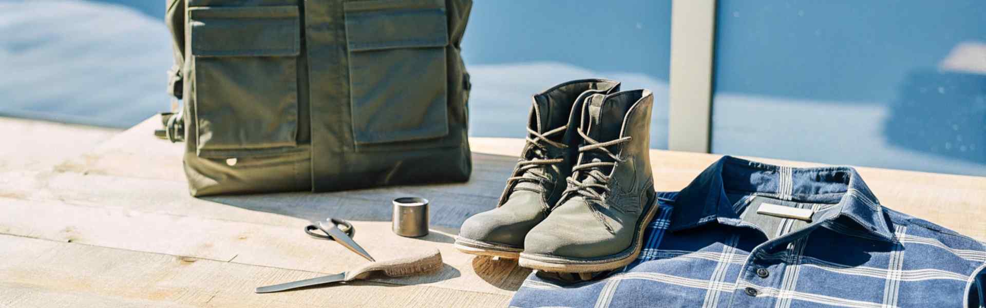 Olive green boots, a green utility bag, and a blue plaid shirt arranged on a wooden table.