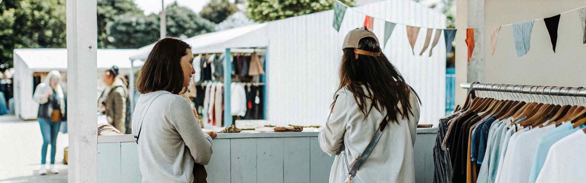 Two women browsing at an outdoor market stall with hanging clothes and bunting decorations.