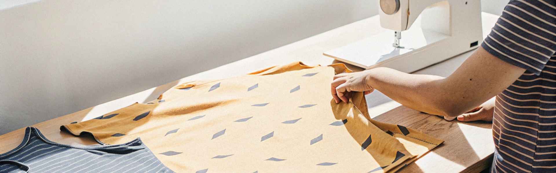 A person adjusting a yellow patterned fabric near a sewing machine on a sunlit table.