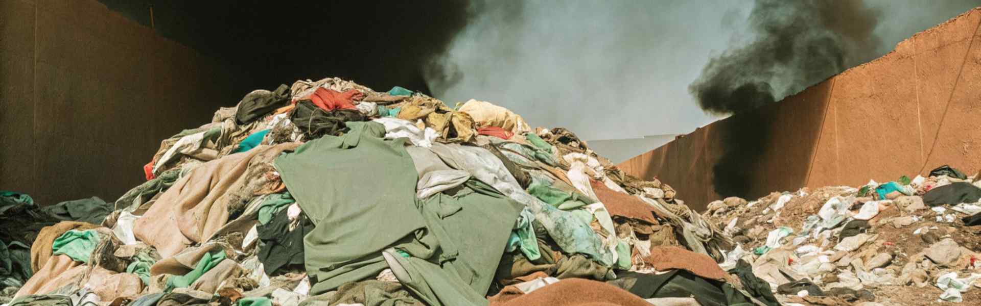 A large pile of discarded clothing in a landfill with black smoke rising in the background.