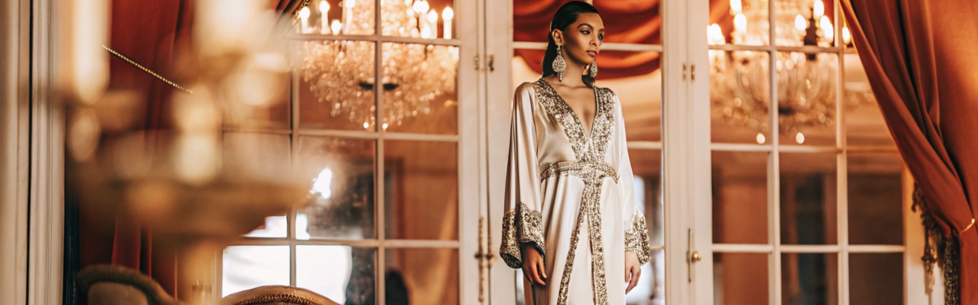 Woman in an ivory gown with intricate gold embellishments stands in a luxurious room with chandeliers and red curtains.