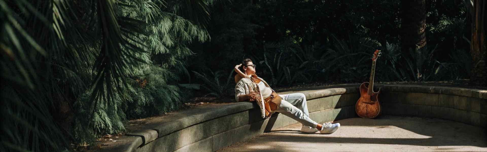 Man reclining on a bench in a lush forest with a guitar resting nearby.