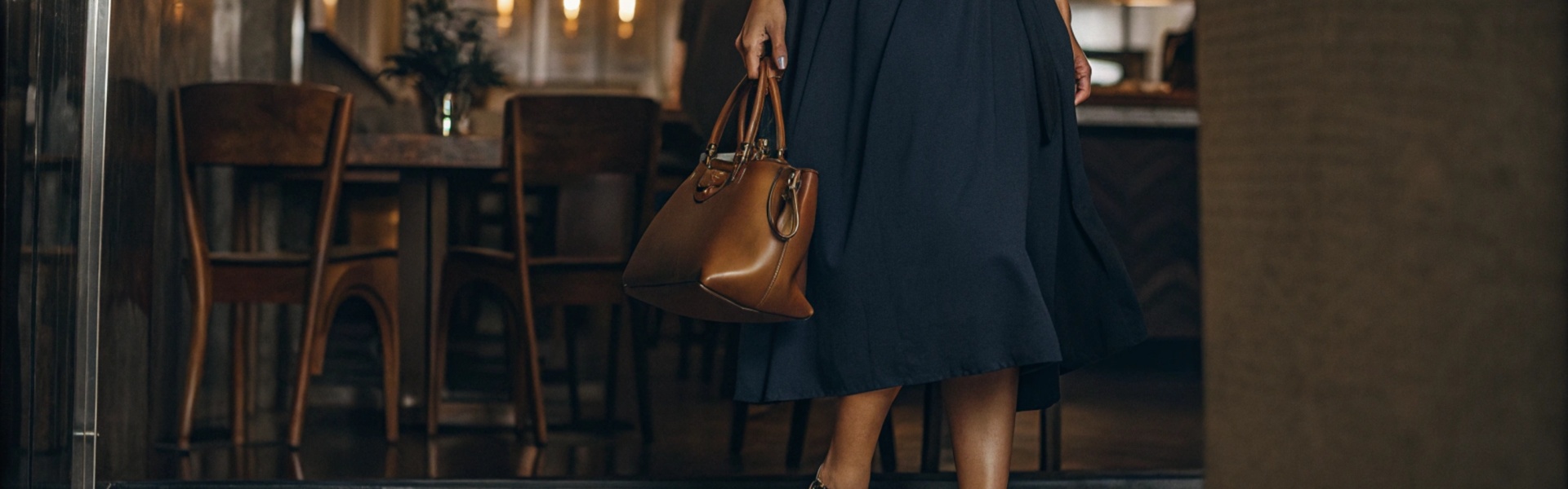 Woman in a navy midi dress with black heels and a tan leather handbag in a stylish cafe setting.