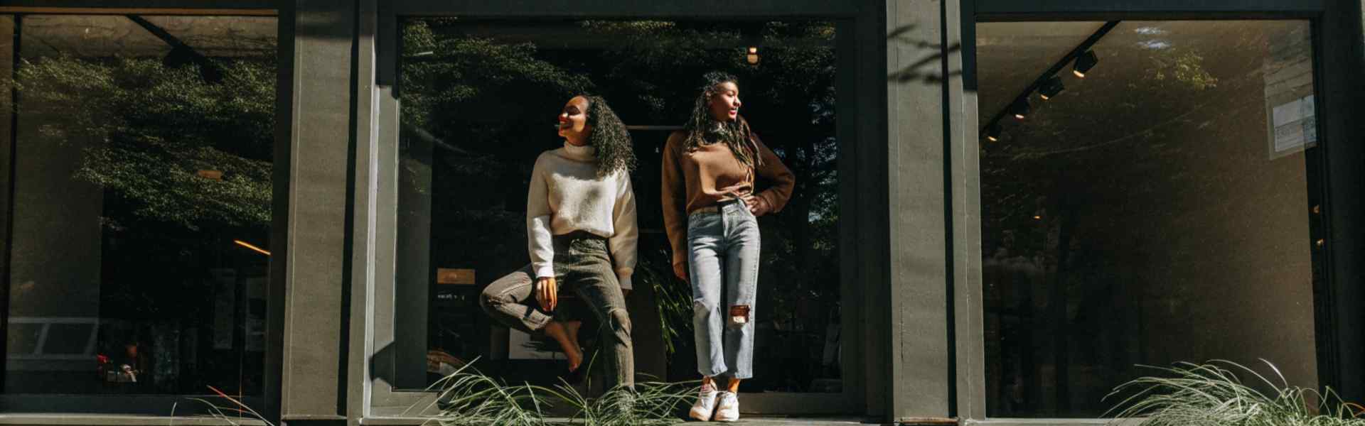 Two women posing by a storefront window surrounded by greenery, showcasing casual outfits.