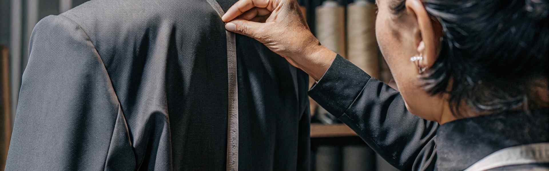 A tailor measuring the back of a black suit jacket using a tape measure.