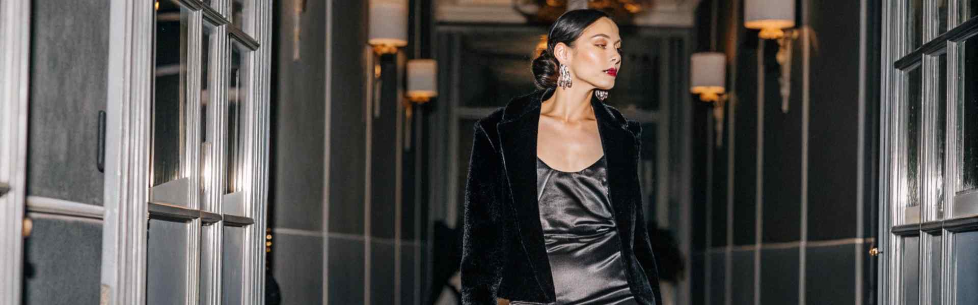 Woman in a black velvet jacket over a satin gown walking down a luxurious hallway.