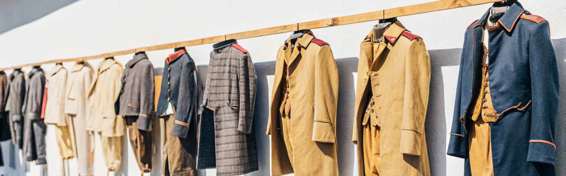 A lineup of vintage-style military and formal coats displayed outdoors on hangers.