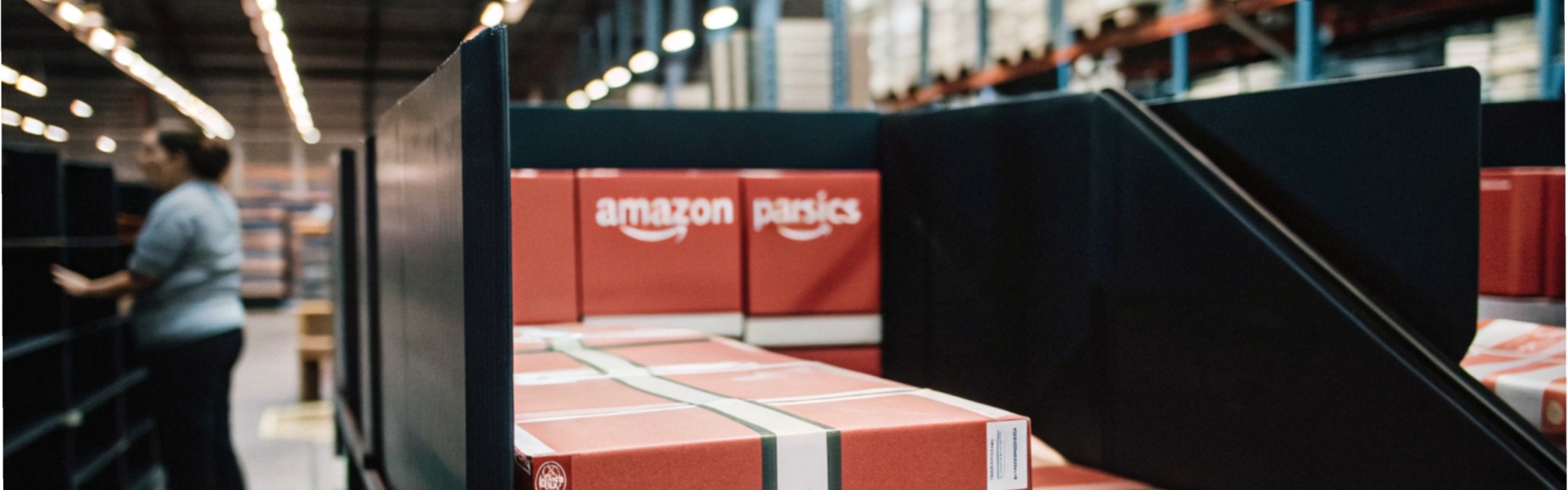 Stacks of red Amazon boxes in a warehouse with shelves and a worker in the background.