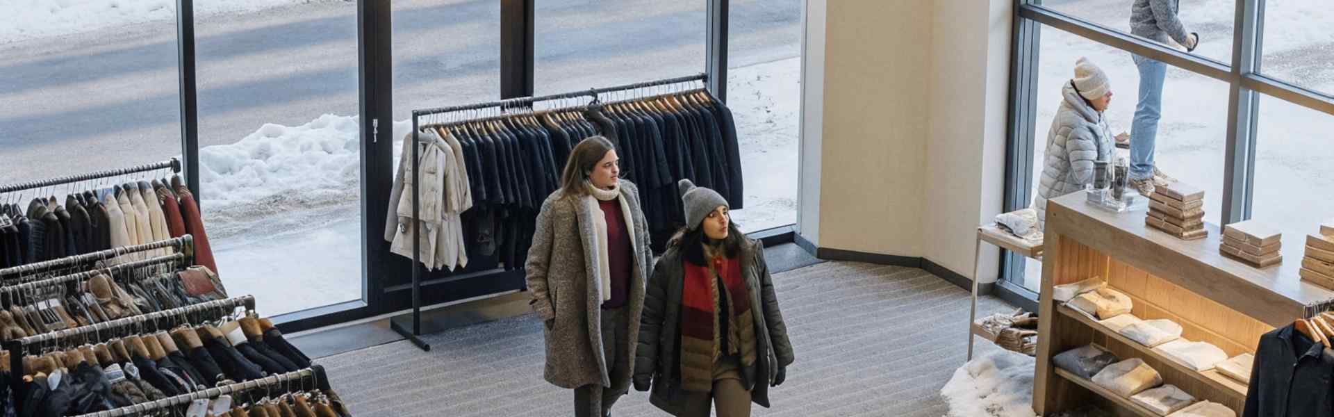 Two women dressed in warm winter coats shopping inside a clothing store with snow visible outside.