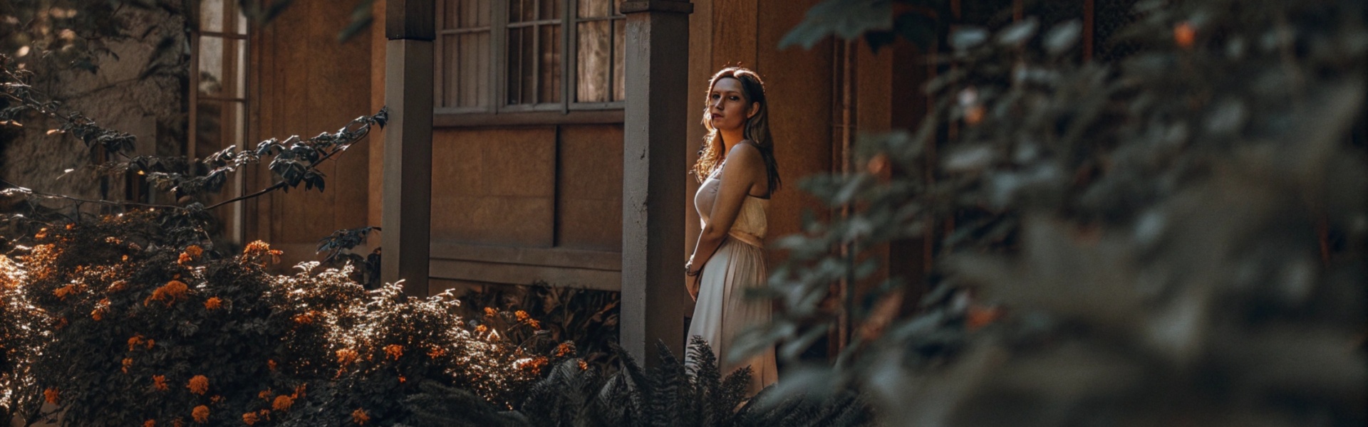 A woman in a flowing white dress stands beside a rustic house, surrounded by vibrant flowers and greenery.