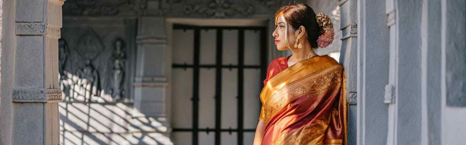 A woman in a red and gold saree standing in a temple corridor, adorned with traditional jewelry and a floral hairstyle.