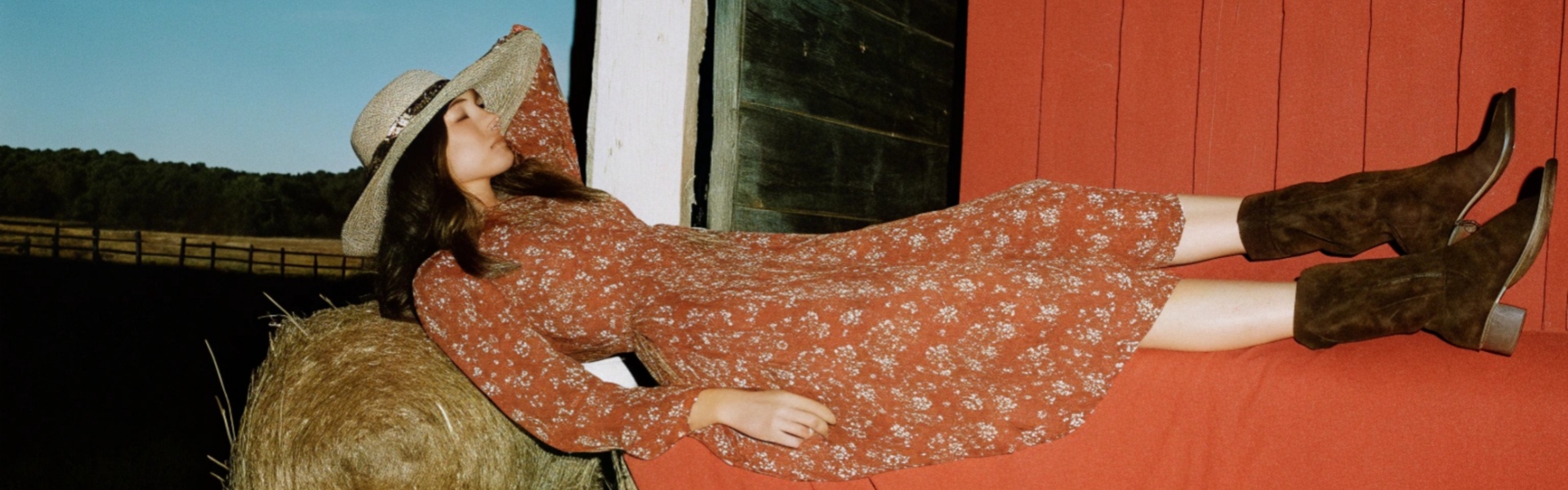 Woman in a floral red dress and brown boots reclining on hay bales near a rustic barn.