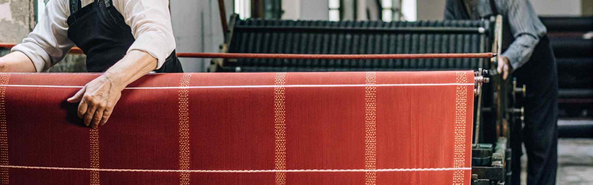 Two craftsmen working with a red fabric roll on traditional textile machinery.