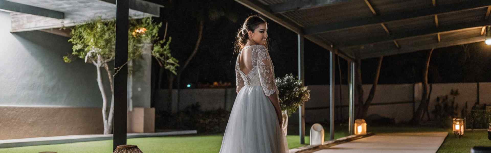 Bride in a lace and tulle gown standing under a covered outdoor walkway with glowing lanterns at night.