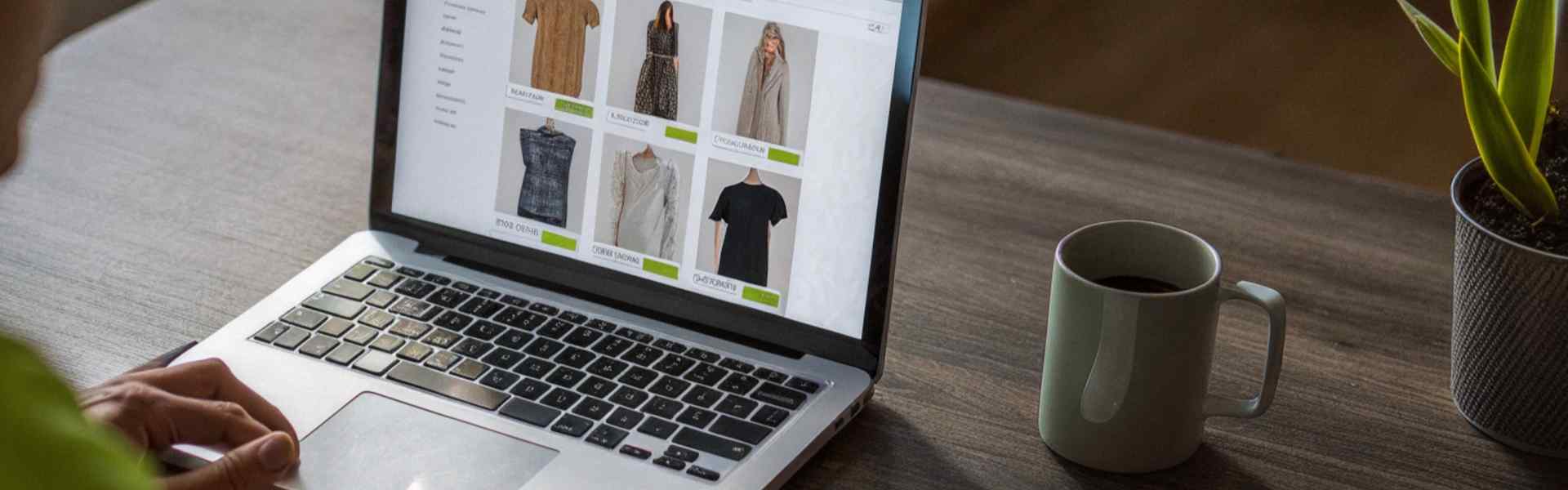 A person browsing a clothing e-commerce site on a laptop, with a notebook, coffee mug, and plant nearby.