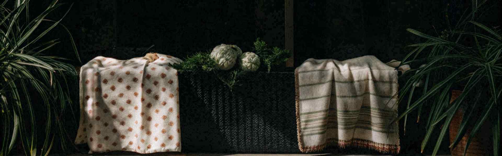 Two patterned blankets draped over a black woven basket surrounded by green plants in soft lighting.