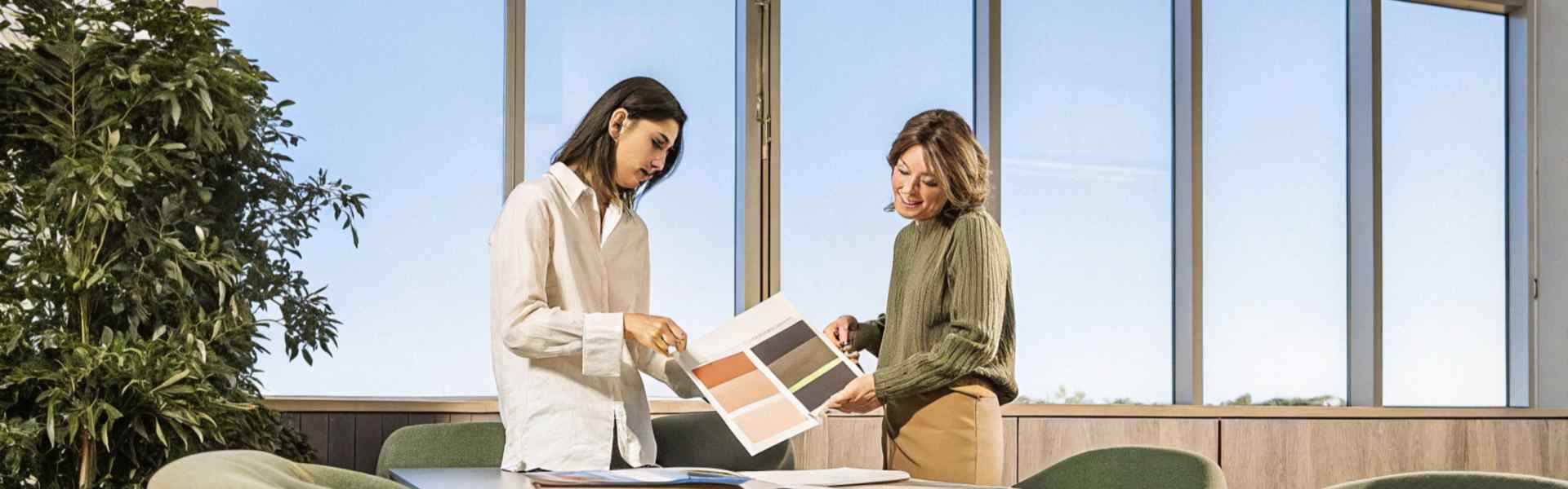 Two women examining a color palette booklet in a well-lit conference room with green chairs and large windows.