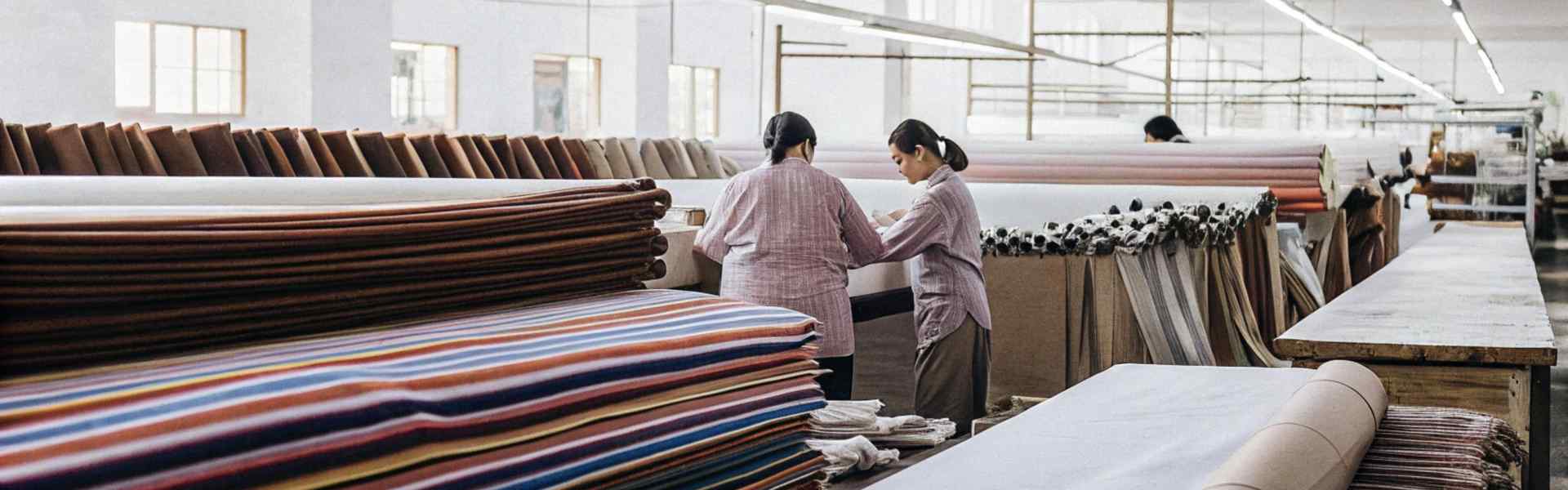 Two women examining colorful fabric rolls in a bright textile factory.