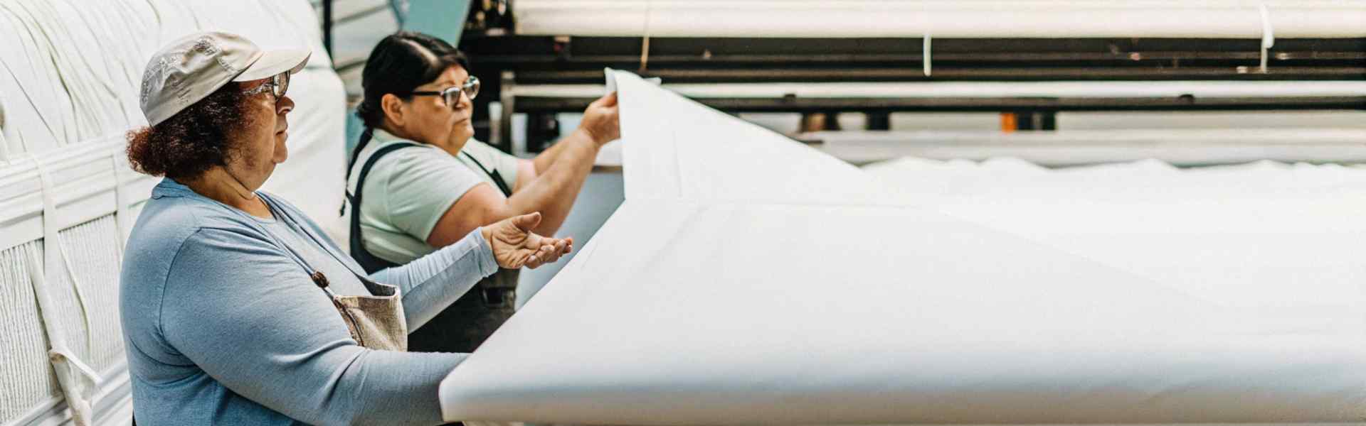 Two workers in a factory inspecting large white fabric rolls during production.