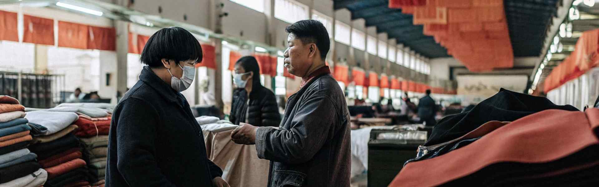 Two individuals wearing masks discussing fabric choices in a large, well-lit indoor market with stacks of colorful textiles.