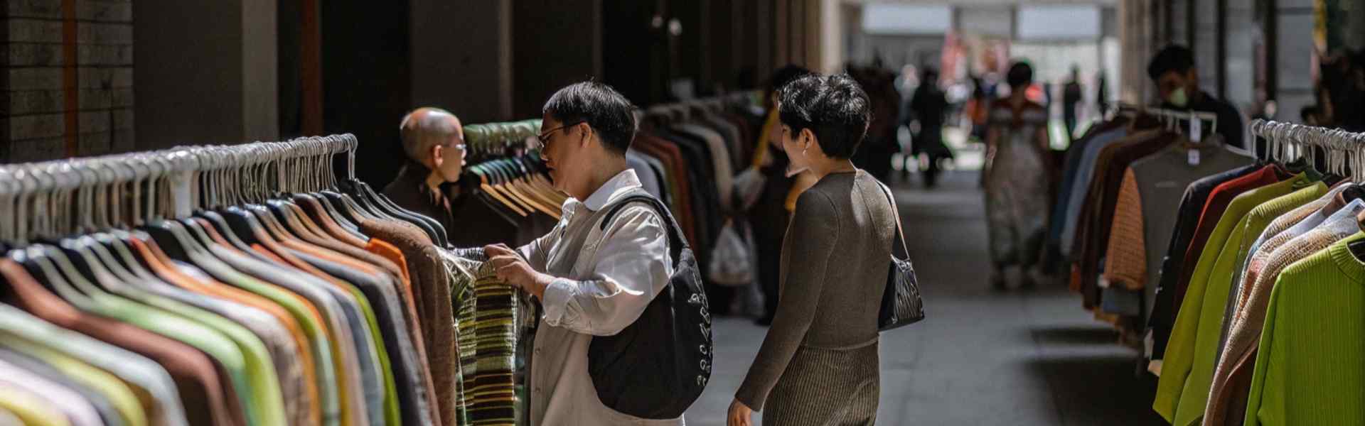 Shoppers browsing colorful garments displayed on racks in a shaded indoor market.