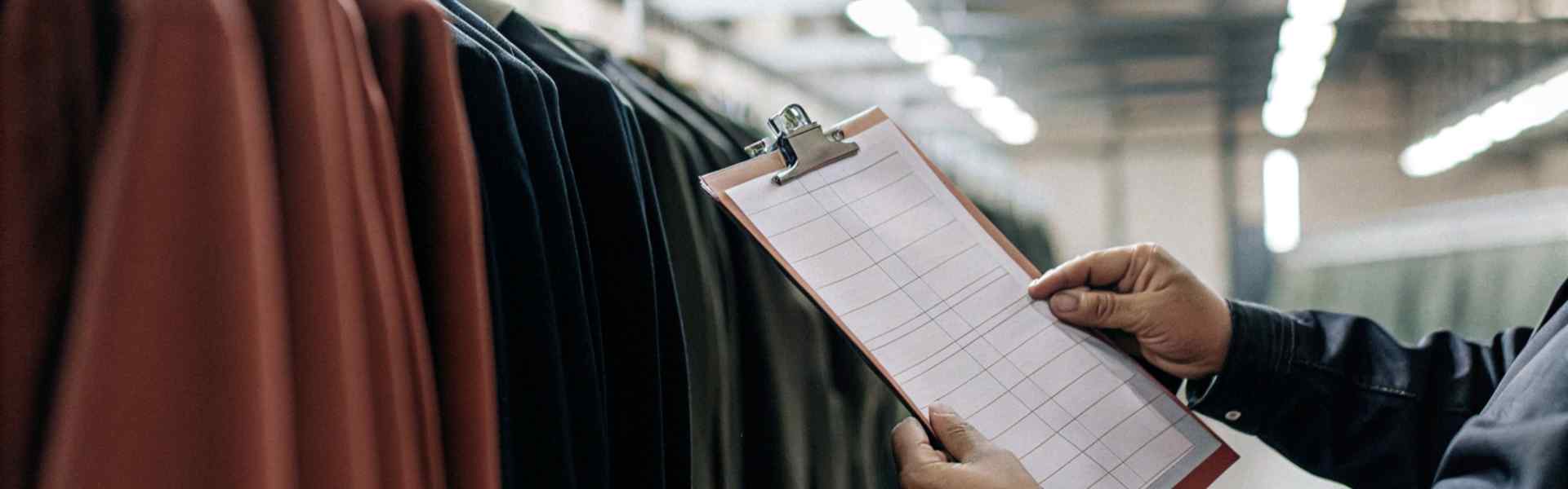 Worker holding a clipboard inspecting clothing racks in a well-lit warehouse.