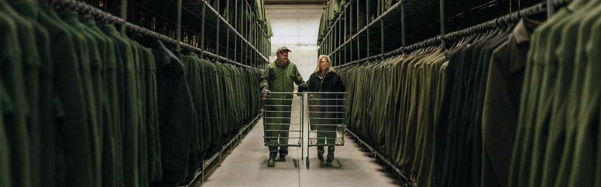 Two workers in green overalls push carts through rows of neatly arranged green uniforms in a storage facility.