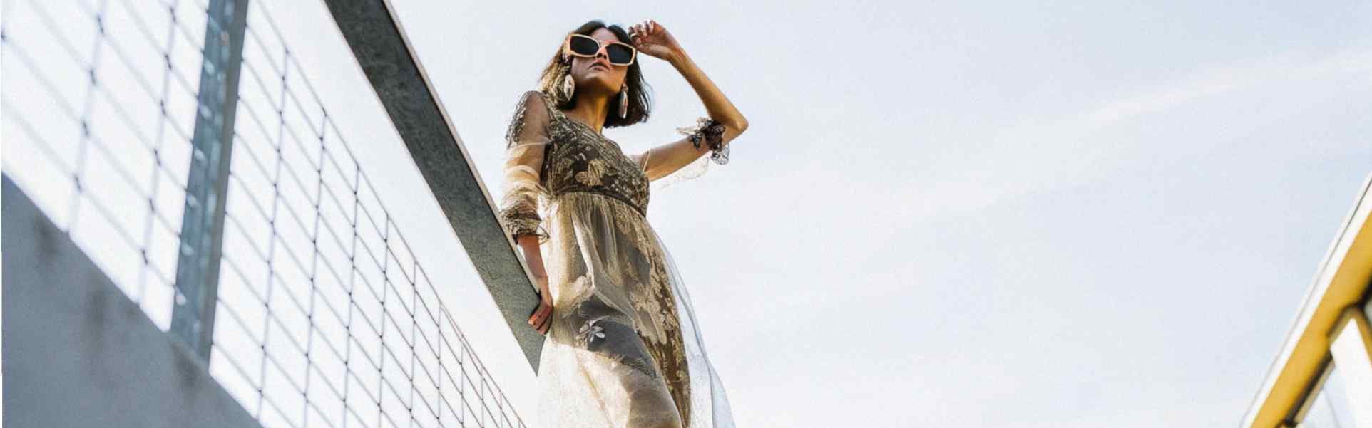 A stylish woman wearing a sheer gold dress and sunglasses poses confidently on a railing under clear skies.