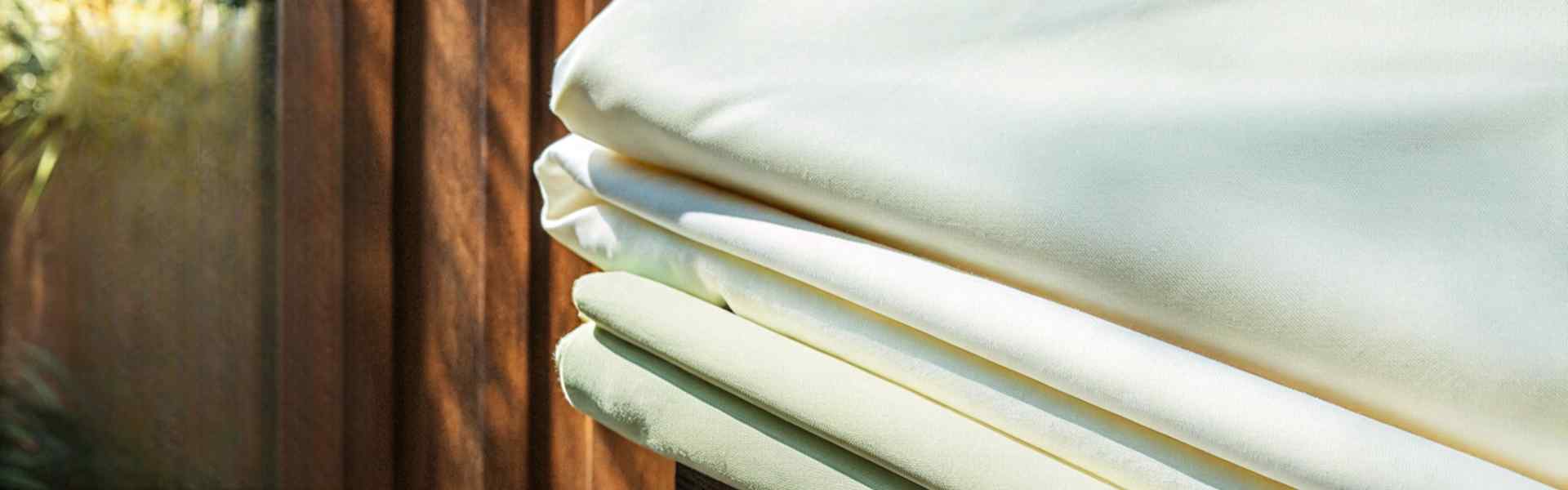 Stacks of cream-colored premium cotton fabric with soft folds, highlighted by sunlight against a wooden background.