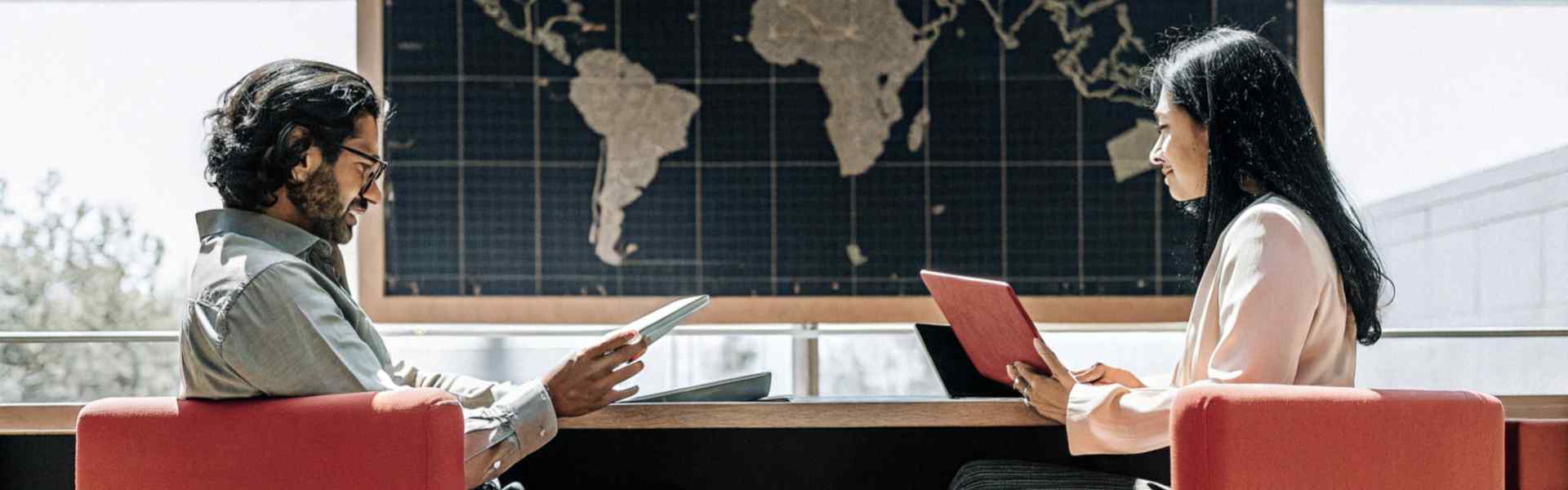 Two professionals sitting on red chairs, engaged in a discussion with tablets, a large world map in the background.