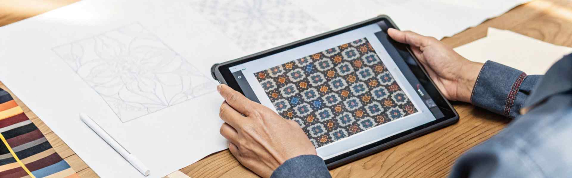 Person viewing intricate fabric patterns on a tablet alongside design sketches on a wooden table.