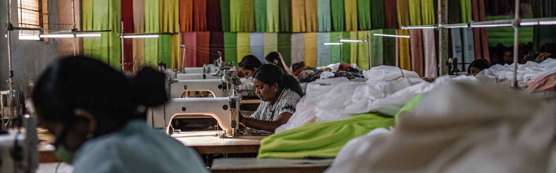 Workers sewing garments in a workshop with colorful fabric rolls hanging in the background.