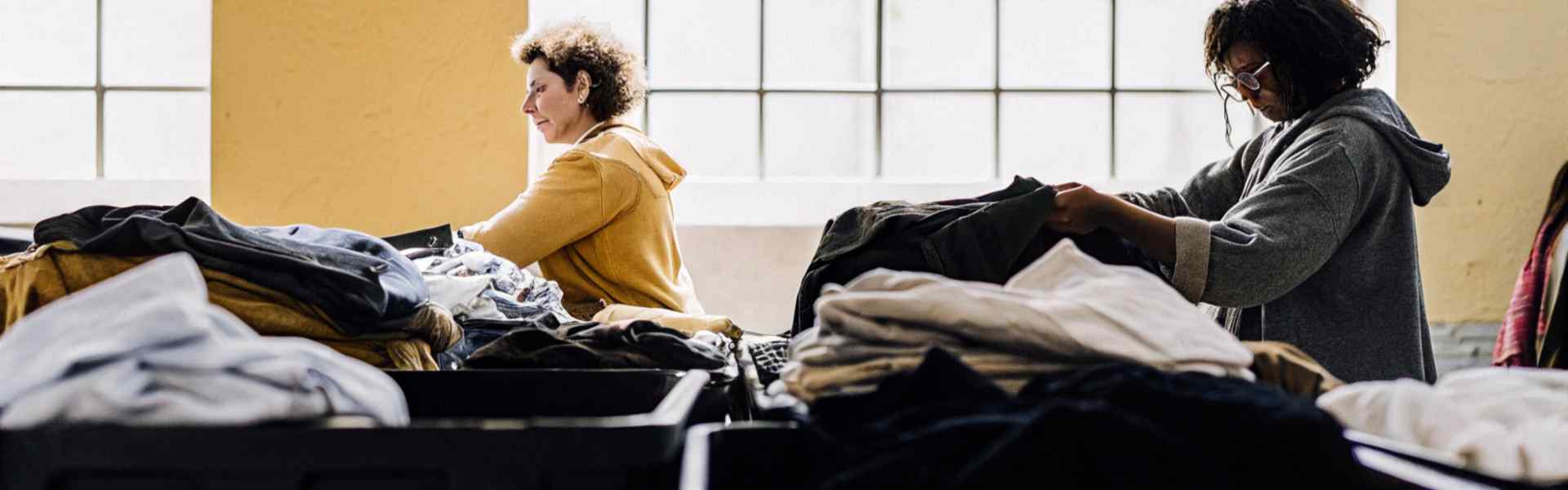 Two women sorting through large piles of clothing in a brightly lit room.