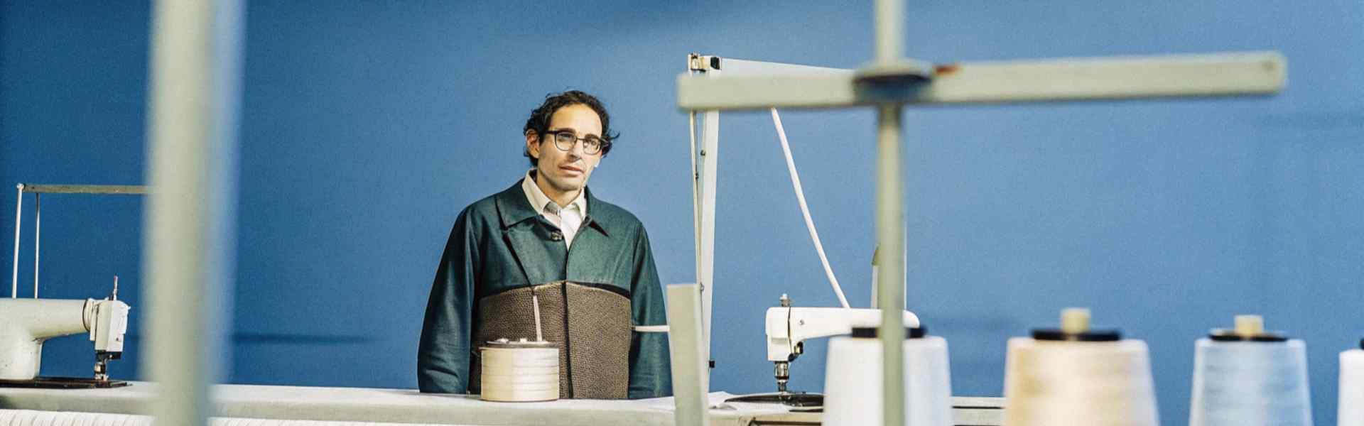 A man standing near sewing machines and spools of thread in a blue-walled textile workspace.