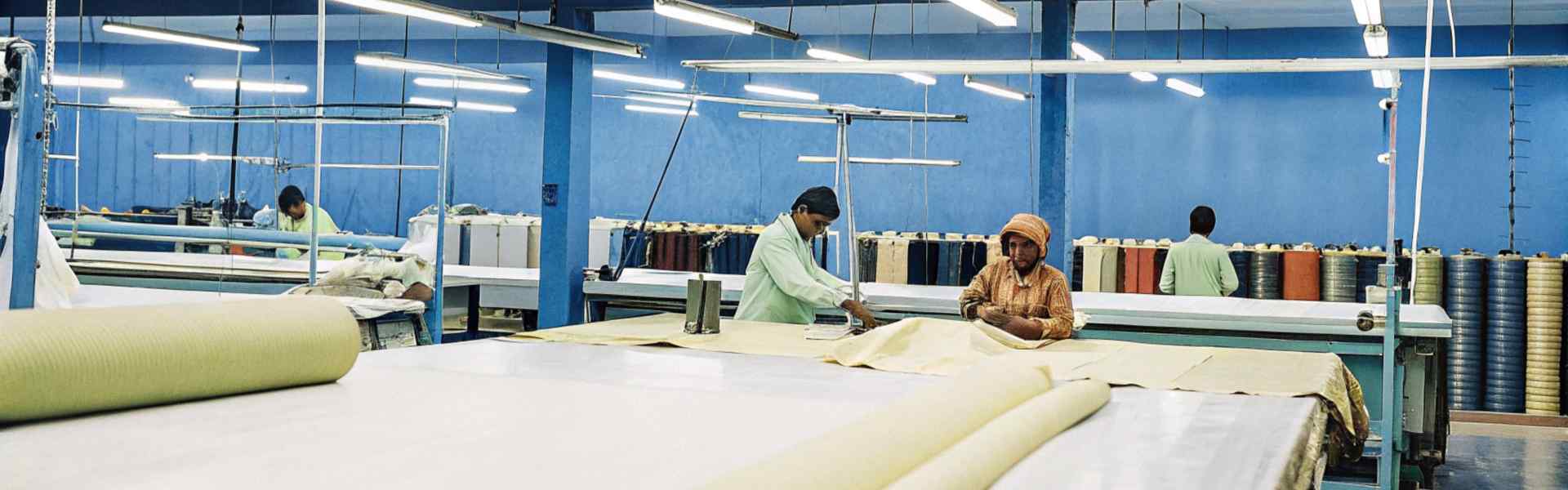 Workers in a brightly lit textile factory cutting and arranging fabric on large tables.