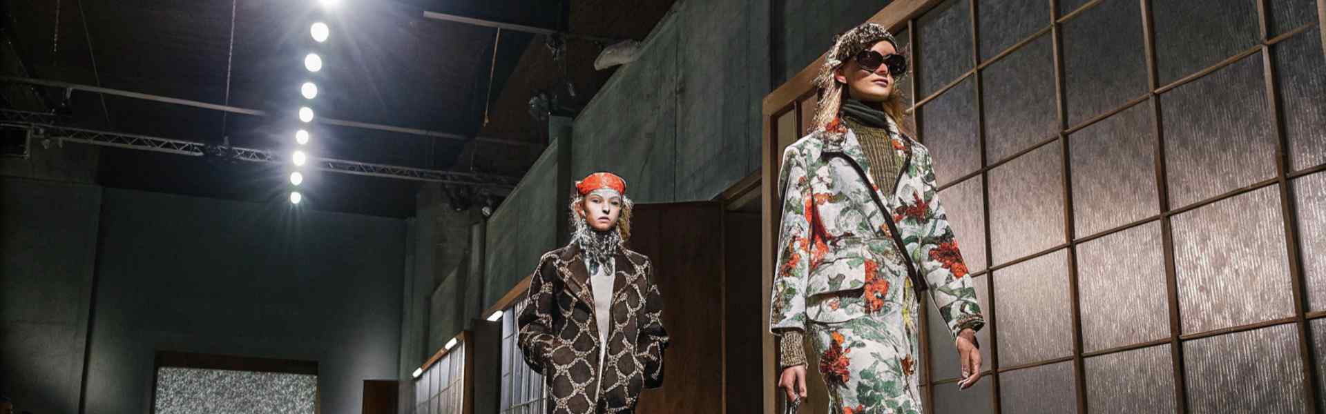 Models in bold patterned outfits walking down a modern runway with green flooring.