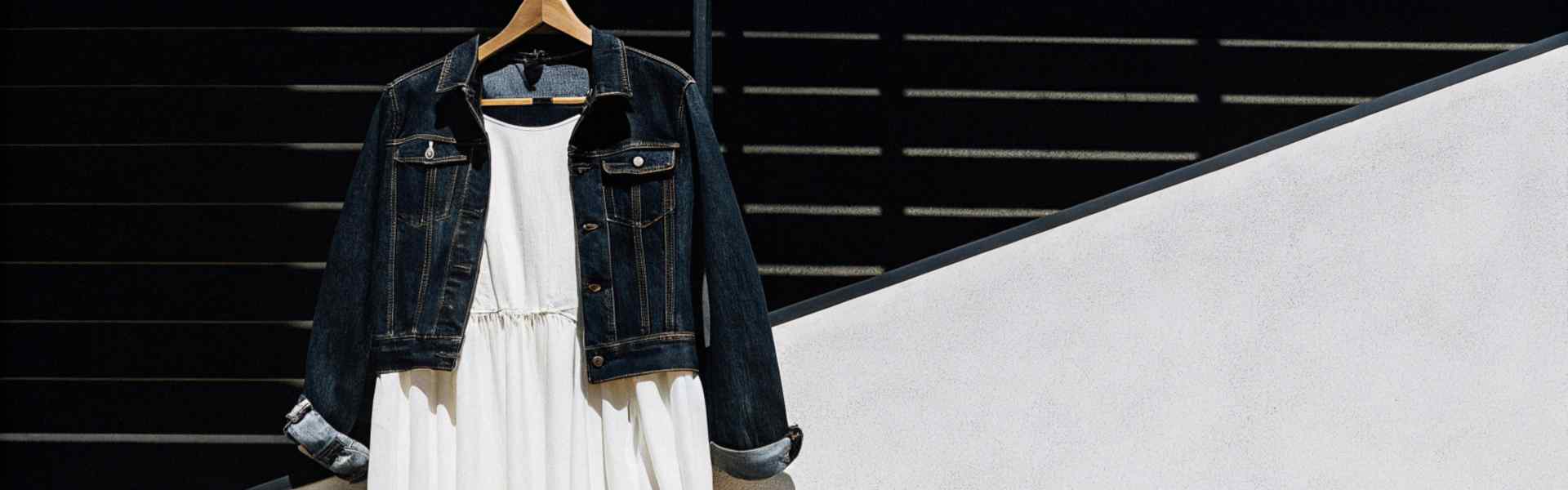 A casual outfit with a white tiered dress paired with a dark denim jacket and white sneakers on a wooden bench.