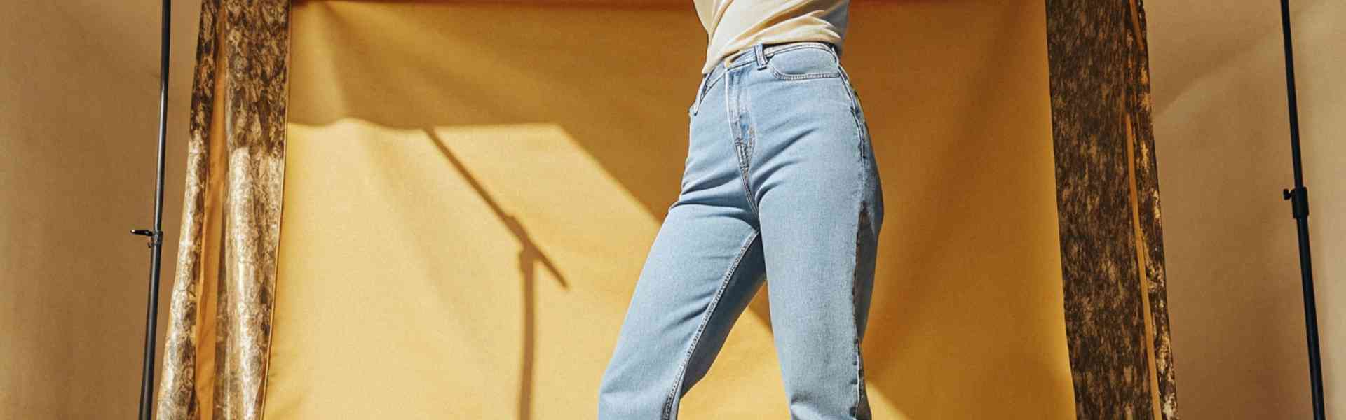 A model wearing high-waisted blue jeans poses in a studio with a golden backdrop.